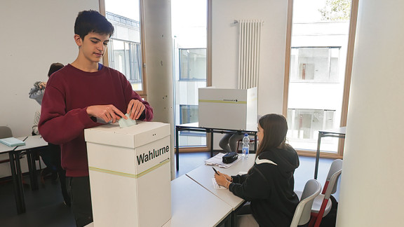 Ein Schüler wirft seinen Stimmzettel in eine Wahlurne. Eine Schülerin sitzt als Wahlhelferin daneben.